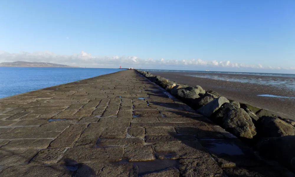Long stone pier extending into the sea with a red lighthouse at the end under a clear blue sky