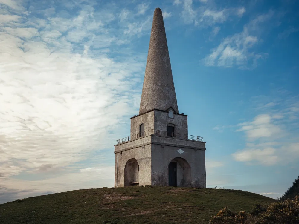 Stone tower on Killiney Hill with arched entrance and viewing platform, surrounded by grassy landscape under a partly cloudy sky
