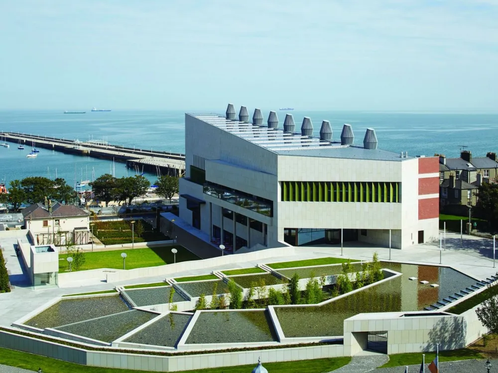 Modern waterfront building with angular architecture, landscaped water features, and nearby boats docked along the pier